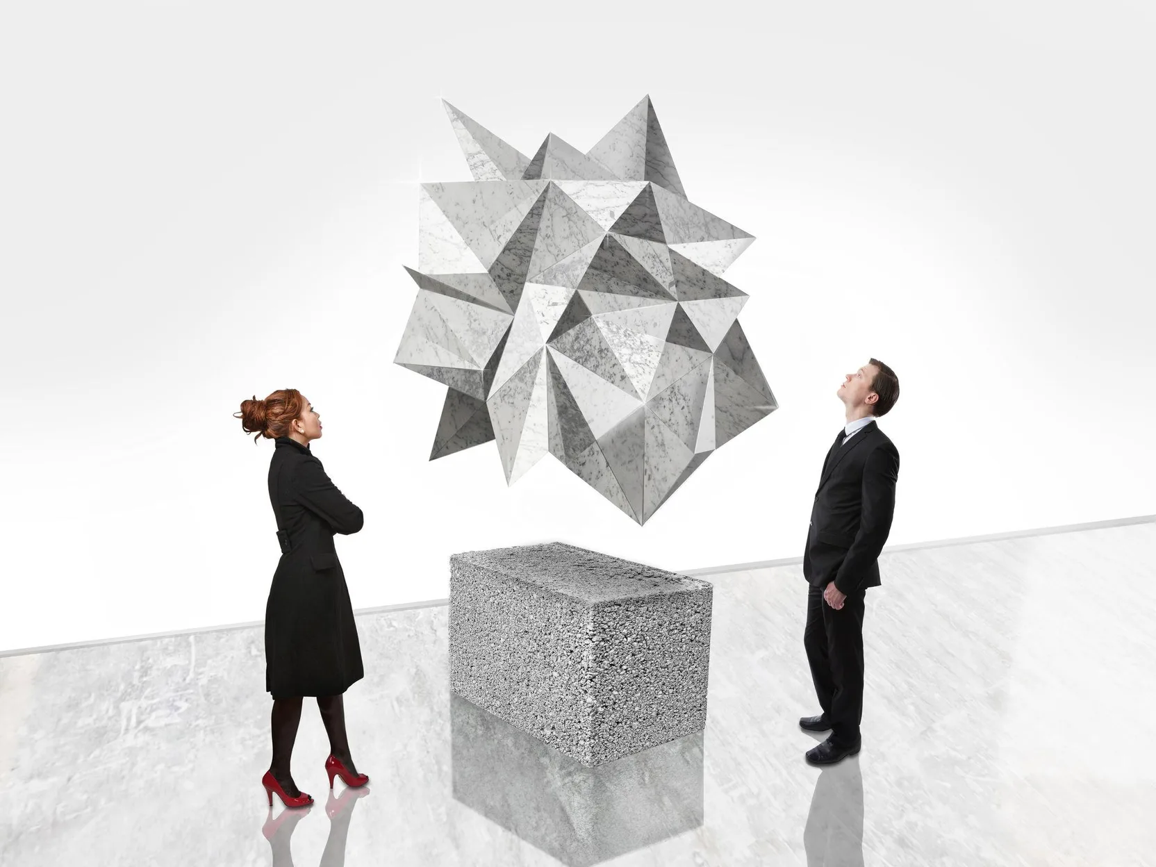 Two people stand on either side of a faceted marble sculpture suspended above a concrete block in a white gallery, each looking up at it from their own position